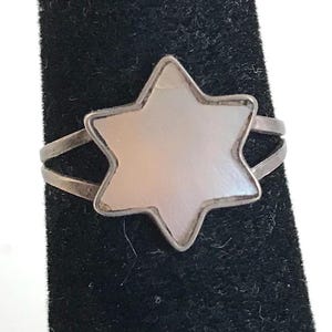 May include: A silver ring featuring a six-pointed star design. The star has a pearlescent, light pink center and a silver border. The ring band splits into two parallel lines on each side of the star.
