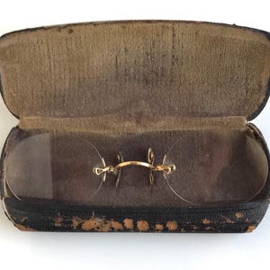 May include: Antique rimless eyeglasses with gold-tone metal bridge and temples, resting inside an open, worn, black leather case with a brown interior. The lenses are clear and oval-shaped.