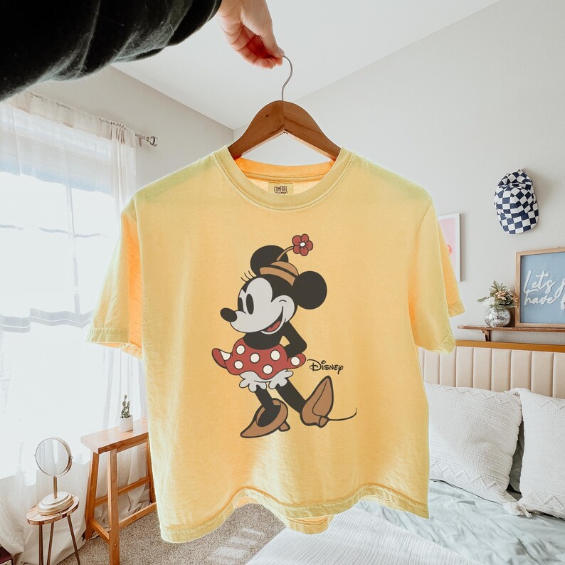 Op de afbeelding: Geel T-shirt met een Minnie Mouse-afbeelding. Het stripfiguur draagt een rood-witte polkadot rok en een hoed met een bloem. Het Disney-logo staat eronder gedrukt.