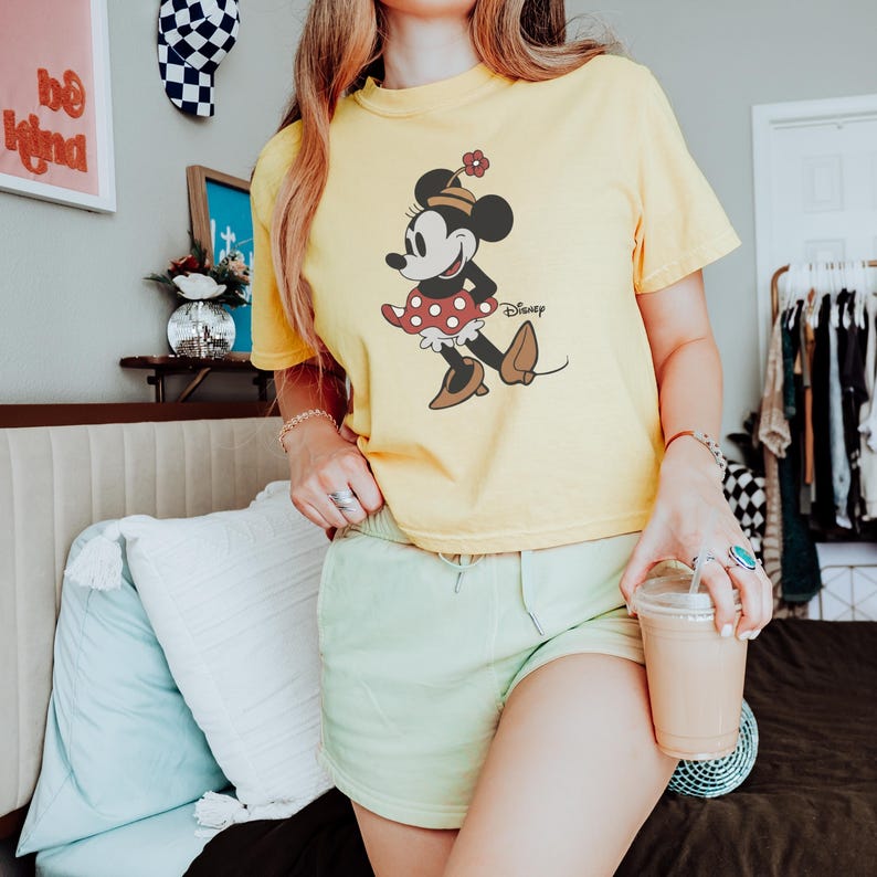 Op de afbeelding: Geel T-shirt met een Minnie Mouse-afbeelding. Het stripfiguur draagt een rode polkadot rok, bruine schoenen en een bloem op haar hoed. Het woord "Disney" staat eronder gedrukt.