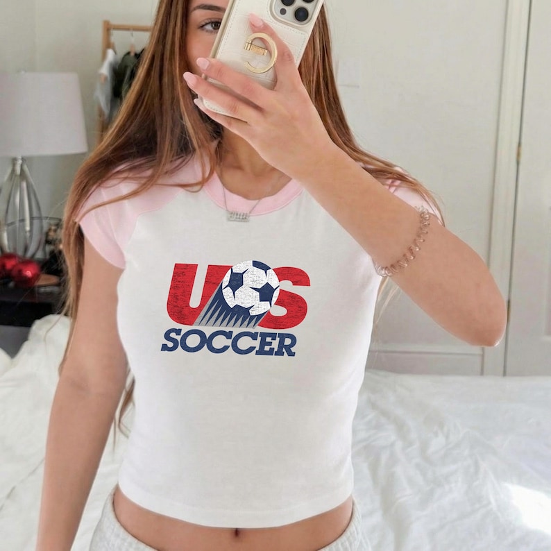 May include: A white short-sleeved ringer t-shirt with light pink trim at the collar and sleeves. The front features a graphic with the red text US, a black and white soccer ball, and the blue text SOCCER printed below the ball graphic.