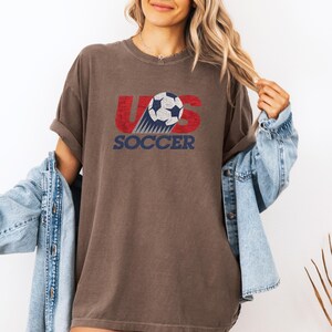 Pode incluir: Uma t-shirt castanha de manga curta com um gr&aacute;fico desgastado em vermelho e azul que diz US SOCCER, com uma bola de futebol a substituir a letra S. A t-shirt &eacute; usada com um casaco de ganga azul claro por cima, num estilo casual.