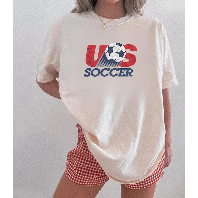 May include: An off-white short-sleeve t-shirt featuring a graphic print on the chest. The design includes the red text US, a black and white soccer ball, and the blue text SOCCER below. The graphic has a distressed, weathered texture.