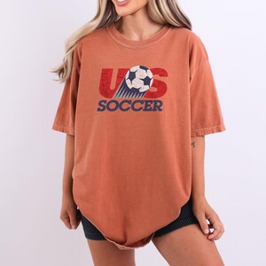 Pode incluir: Uma t-shirt de manga curta cor de ferrugem com uma impress&atilde;o gr&aacute;fica no peito. O design inclui o texto vermelho US com uma bola de futebol a substituir a letra S, e a palavra SOCCER em azul por baixo, tudo com uma textura desgastada.