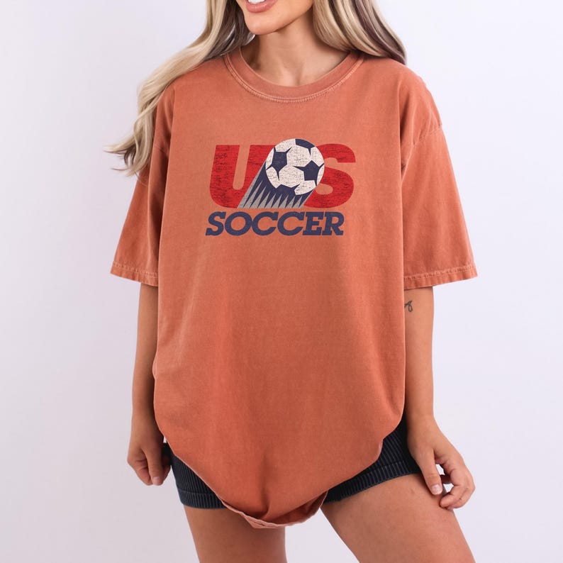 Pode incluir: Uma t-shirt de manga curta cor de ferrugem com uma impress&atilde;o gr&aacute;fica no peito. O design inclui o texto vermelho US com uma bola de futebol a substituir a letra S, e a palavra SOCCER em azul por baixo, tudo com uma textura desgastada.