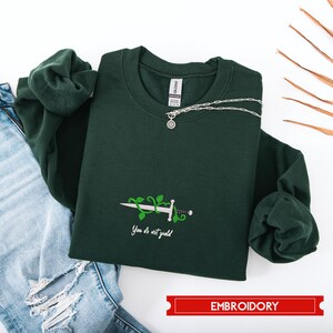 May include: A dark green sweatshirt with a crew neck, featuring an embroidered design of a sword entwined with green vines and the text "You do not yield." A silver necklace is draped across the sweatshirt.