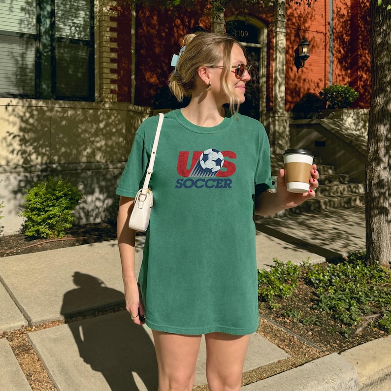 Pode incluir: Uma t-shirt de manga curta verde-floresta com um gr&aacute;fico no peito com o texto US SOCCER em vermelho e azul, acompanhado por uma ilustra&ccedil;&atilde;o de uma bola de futebol. A camisa tem gola redonda e um corte descontra&iacute;do, com uma mala de ombro branca.