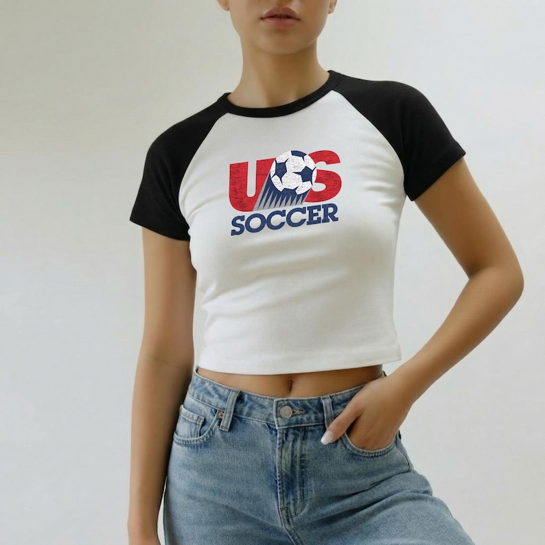 May include: A white and black raglan t-shirt featuring a graphic of a soccer ball and the red text US with the blue text SOCCER underneath. The design includes a motion trail effect behind the ball, creating a dynamic look on the front of the shirt.
