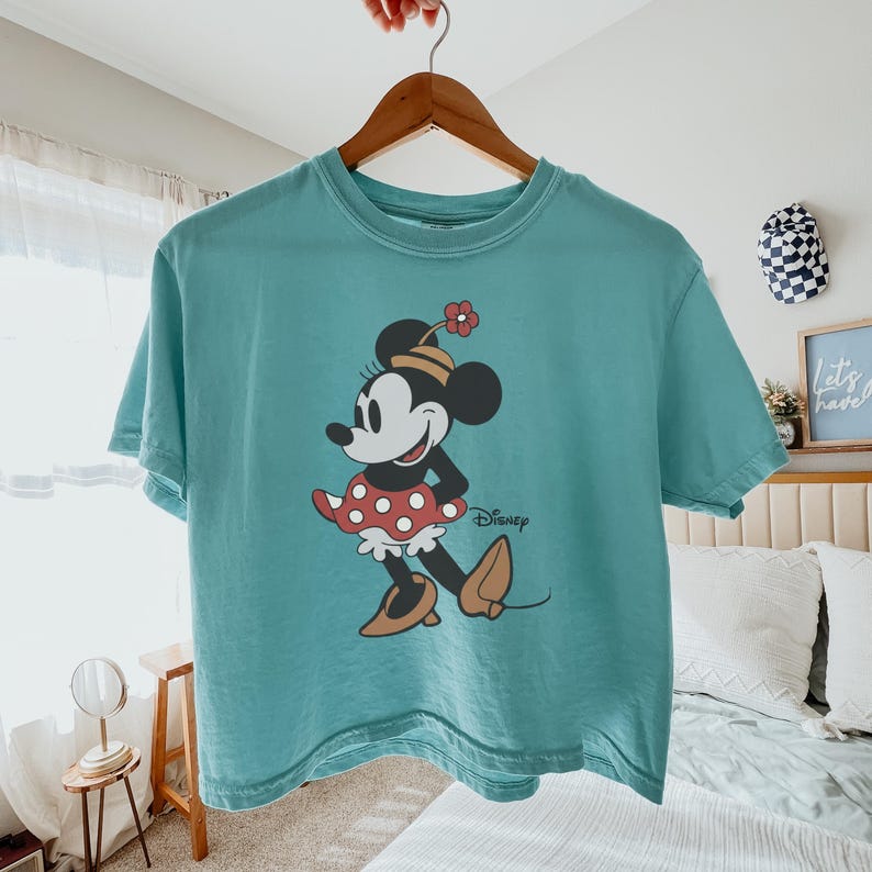 Op de afbeelding: Een turquoise t-shirt met een Minnie Mouse-afbeelding. Het stripfiguur is afgebeeld in een rood-witte polkadot rok, een bloemenhoed en bruine schoenen. Het Disney-logo staat eronder.
