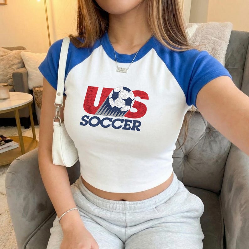 May include: A white short-sleeve raglan t-shirt with blue sleeves and a graphic on the chest. The graphic features the red text US with a soccer ball replacing the letter O, and the word SOCCER printed in blue underneath the graphic.