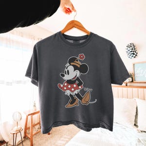 Op de afbeelding: Donkergrijs t-shirt met een Minnie Mouse-afbeelding. Minnie wordt afgebeeld in een rode polkadotjurk, bruine schoenen en een hoed met een bloem. Het Disney-logo staat eronder.