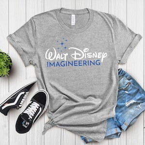 May include: A heather gray t-shirt with the text "Walt Disney Imagineering" in white and blue. The shirt is paired with black and white sneakers and denim shorts. The shirt is made of a soft material.