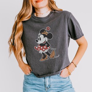 Op de afbeelding: Donkergrijs T-shirt met een Minnie Mouse-afbeelding. Het stripfiguur draagt een rode jurk met witte stippen, bruine schoenen en een bloem op haar hoed. Het Disney-logo staat op het shirt.