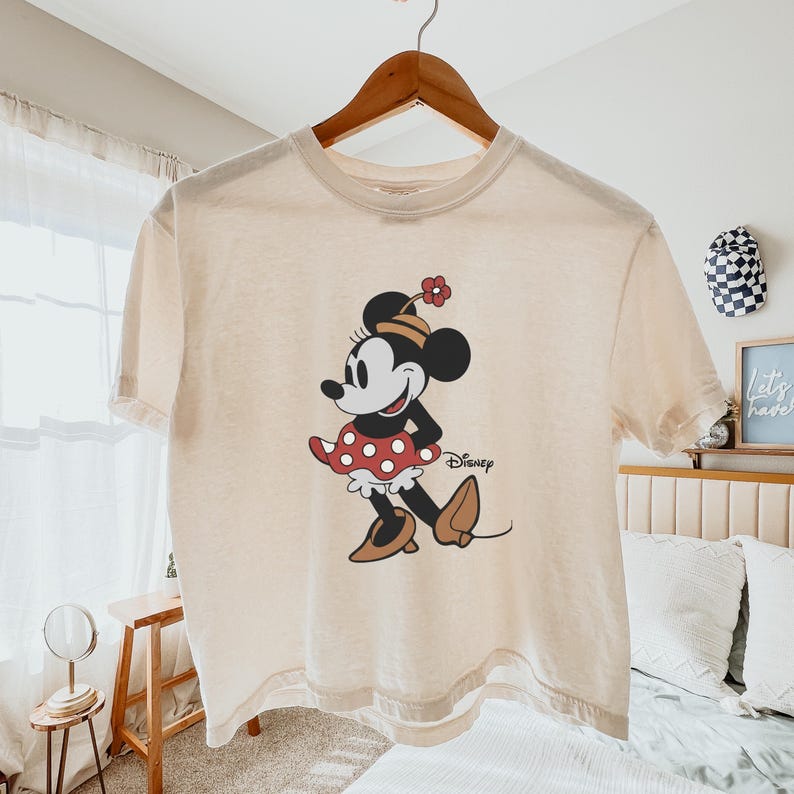 Op de afbeelding: Cr&egrave;mekleurig T-shirt met een klassieke Minnie Mouse afbeelding. Minnie is afgebeeld in haar iconische rode polkadotjurk, hoed en schoenen. Het Disney-logo staat eronder. Het shirt hangt aan een houten hanger.