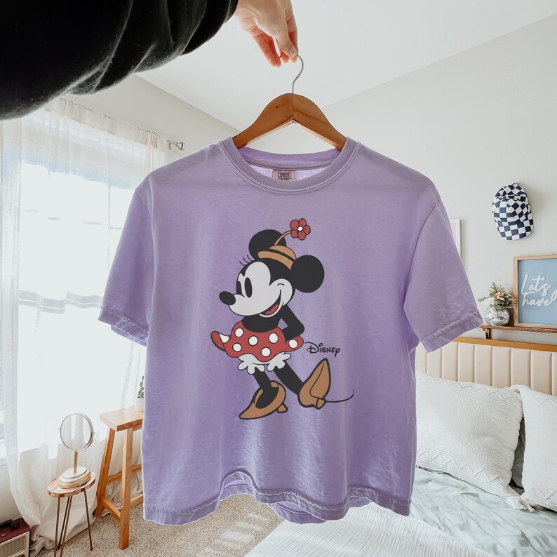 Op de afbeelding: Een lavendelkleurig T-shirt met een afbeelding van Minnie Mouse. Het stripfiguur draagt een rode polkadot rok, een bloemenhoed en bruine schoenen. Het woord "Disney" staat eronder.