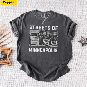 May include: Dark gray t-shirt with the text "STREETS OF MINNEAPOLIS" and a black and white image of a protest. The image includes signs that say "I THOUGHT ALL LIVES MATTER" and "ICE OUT".