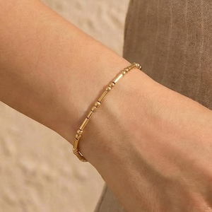 May include: A gold bracelet featuring a series of small, cylindrical beads and rounded accents. The bracelet is worn on a wrist, showcasing its delicate design and warm, metallic color. The jewelry piece is a simple yet elegant accessory.
