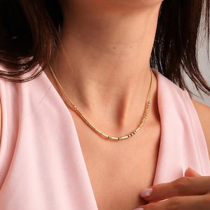 Könnte beinhalten: Eine filigrane goldene Halskette mit kleinen, gleichmäßig verteilten Perlen und zylindrischen Stäben. Die Kette liegt am Hals einer Person. Der Schmuck wird vor einem zarten rosa Hintergrund präsentiert.