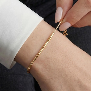May include: A gold bracelet featuring a series of small beads and cylindrical sections. The bracelet is being adjusted on a wrist, highlighting its adjustable design. The jewellery is simple and elegant, suitable for everyday wear.