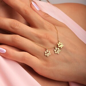 May include: A gold-colored necklace featuring three paw print pendants, each inscribed with a name. The necklace is displayed on a person's hand, with a pink background. The pendants are detailed with small paw pad impressions.