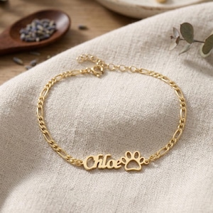 May include: A gold-coloured bracelet with a chain link design. The bracelet features the name "Chloe" and a paw print charm. The jewellery is displayed on a neutral-coloured cloth, with a wooden spoon and other jewellery in the background.