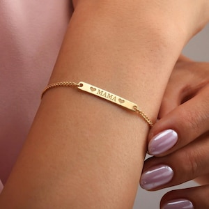 May include: A gold-colored bracelet with a delicate chain and a rectangular bar. The bar is engraved with the word "MAMA" between two heart symbols. The bracelet is worn on a wrist.