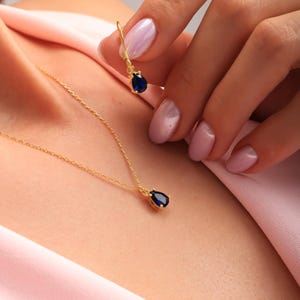 May include: A gold necklace and earring set featuring teardrop-shaped, dark blue gemstones. The necklace is worn around the neck, and the earring is held up by a hand. The jewelry is set against a pink background.