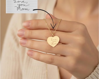May include: A gold-colored heart-shaped pendant necklace. The pendant has the words "Love, you Mom" engraved on it. The necklace is held by a person. A handwritten note with the same text is shown above the necklace.
