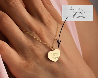 May include: A gold-colored heart pendant necklace. The heart-shaped pendant has the handwritten inscription "Love, you Mom". The necklace is on a delicate chain. A handwritten note with the same inscription is in the background.