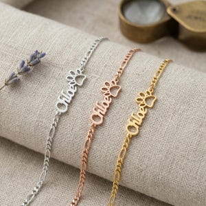 May include: Three personalized bracelets with the name "Chloe" and a paw print charm. The bracelets are silver, rose gold, and gold. The bracelets are displayed on a neutral linen surface.