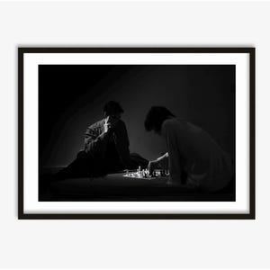 Może przedstawiać: Czarno-biała oprawiona grafika przedstawiająca dwie osoby grające w szachy. Jedna osoba siedzi, opierając podbródek na dłoni, podczas gdy druga pochyla się nad szachownicą, wykonując ruch. Figury szachowe są widoczne.