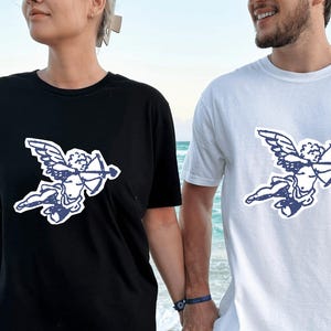 Peut inclure: T-shirts noirs et blancs avec un motif de cupidon bleu. Le cupidon est représenté avec des ailes, un arc et une flèche. Les t-shirts sont portés par deux personnes sur une plage de sable, avec la mer en arrière-plan.