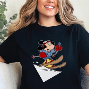 Puede incluir: Camiseta negra con un dibujo animado de Mickey Mouse esquiando. Mickey lleva una chaqueta azul, guantes rojos y gafas. Los esquís son marrones y la nieve es blanca. El diseño es una ilustración colorida.