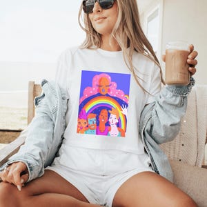 May include: White t-shirt with a colorful graphic of diverse figures under a rainbow. The artwork features a woman with pink hair and a rainbow arching over several smiling faces. The shirt is part of a matching set with white shorts.