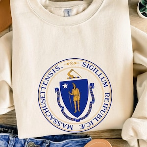 Puede incluir: Sudadera color crema con el sello del estado de Massachusetts. El sello es un diseño circular azul y dorado, con texto alrededor. La sudadera está hecha de un material suave y tiene cuello redondo.