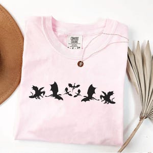 May include: A light pink t-shirt with a crew neck featuring a black dragon silhouette design. The shirt has a small tag that says "Comfort Colors". A silver necklace with a small pendant is draped on the shirt.