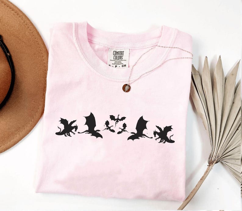 May include: A light pink t-shirt with a crew neck featuring a black dragon silhouette design. The shirt has a small tag that says "Comfort Colors". A silver necklace with a small pendant is draped on the shirt.