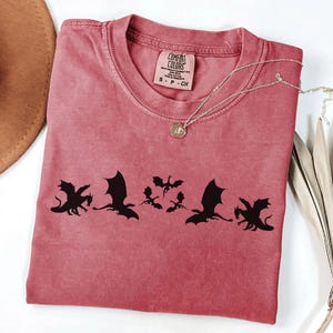 May include: A dusty rose Comfort Colors t-shirt featuring a black dragon silhouette design. The shirt has a crew neck and short sleeves. A gold necklace with a pendant is draped across the shirt. A brown hat and dried plant are in the background.