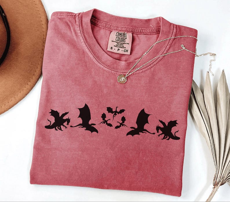 May include: A dusty rose Comfort Colors t-shirt featuring a black dragon silhouette design. The shirt has a crew neck and short sleeves. A gold necklace with a pendant is draped across the shirt. A brown hat and dried plant are in the background.