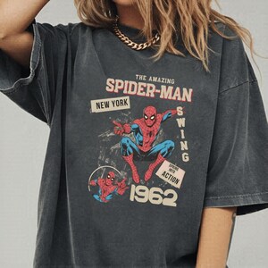 May include: Dark grey t-shirt with a vintage Spider-Man graphic. The design includes the text "THE AMAZING SPIDER-MAN," "NEW YORK," and "1962." The superhero is shown in his classic red and blue suit.