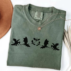 May include: An olive green t-shirt with a black dragon silhouette design. The shirt has a crew neck and short sleeves. A gold necklace with a pendant is draped across the shirt. A brown hat and dried plant are in the background.