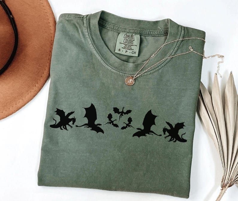 May include: An olive green t-shirt with a black dragon silhouette design. The shirt has a crew neck and short sleeves. A gold necklace with a pendant is draped across the shirt. A brown hat and dried plant are in the background.