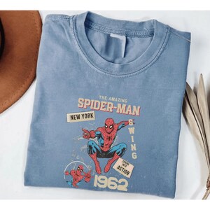 May include: A pale blue t-shirt with a retro Spider-Man design. The graphic includes the text "The Amazing Spider-Man," "New York," and the year "1962." The superhero is shown in his red and blue suit, in a dynamic pose.