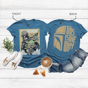 Op de afbeelding: Twee tealkleurige t-shirts met Star Wars-graphics. E&eacute;n toont The Mandalorian en Grogu, de andere heeft een helmdesign en de tekst "THIS IS THE WAY". De shirts worden getoond met witte sneakers, jeans en een koffie.