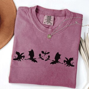 May include: A mauve Comfort Colors t-shirt featuring a black dragon silhouette design. The shirt has a crew neck and short sleeves. A silver necklace with a pendant is draped across the shirt. The tag reads "Comfort Colors".