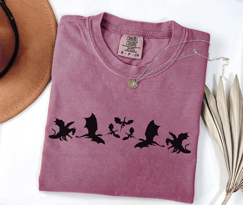 May include: A mauve Comfort Colors t-shirt featuring a black dragon silhouette design. The shirt has a crew neck and short sleeves. A silver necklace with a pendant is draped across the shirt. The tag reads "Comfort Colors".