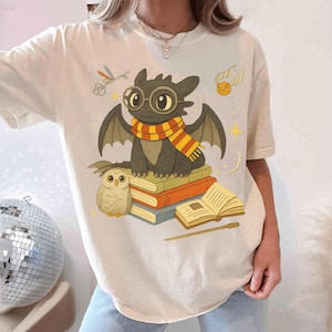 May include: Cream-colored t-shirt featuring a cartoon dragon wearing glasses and a scarf, sitting on books. An owl, a golden snitch, and other magical elements are also depicted. The design is printed on a soft, casual tee.