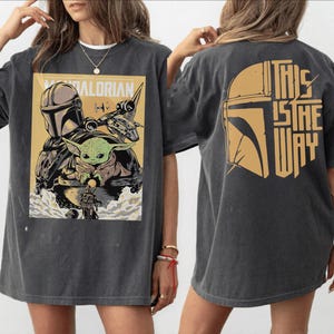 Op de afbeelding: Donkergrijs T-shirt met een afbeelding van The Mandalorian. De voorkant toont de Mandalorian, Grogu en een schip, met tekst. De achterkant heeft een helm-afbeelding en de zin "This is the Way" in goud.