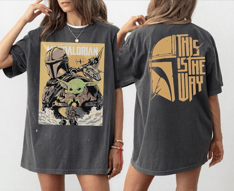 Op de afbeelding: Donkergrijs T-shirt met een afbeelding van The Mandalorian. De voorkant toont de Mandalorian, Grogu en een schip, met tekst. De achterkant heeft een helm-afbeelding en de zin "This is the Way" in goud.