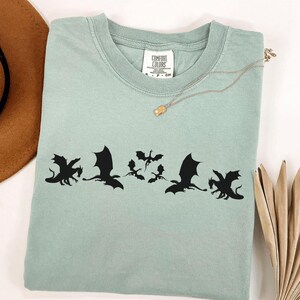 May include: A light green t-shirt with a black dragon graphic. The shirt has a crew neck and short sleeves. A gold necklace with a small pendant is on the shirt. A brown hat and dried plant are in the background.